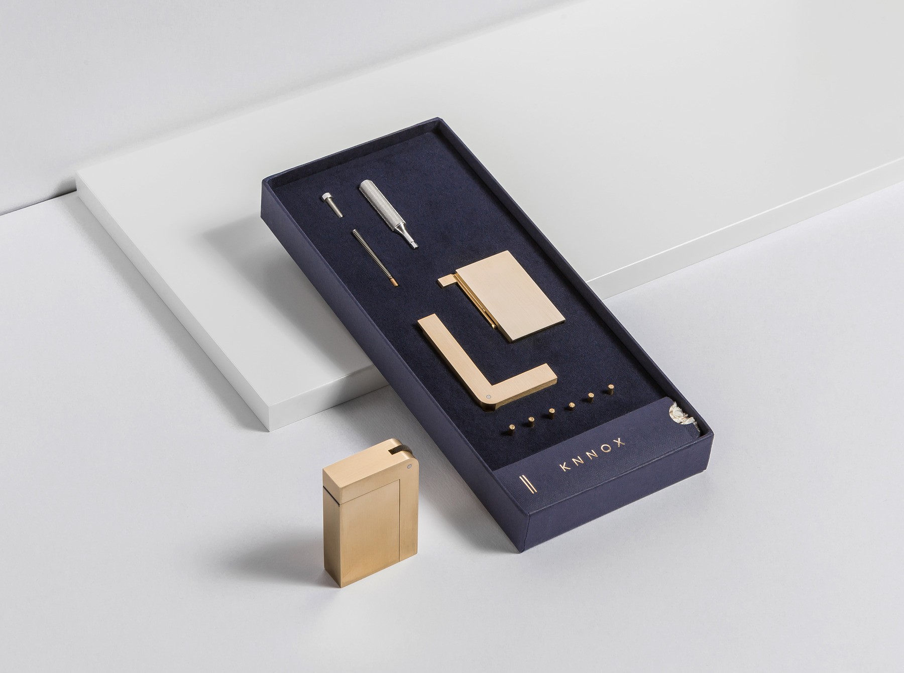 A brass-toned KNNOX object stands beside a dark blue tray containing carefully arranged components on a white studio set.